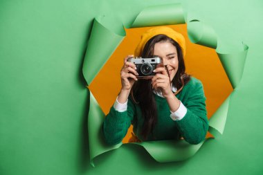 Young white smiling woman peeking out hole isolated on green background taking photos with camera clipart