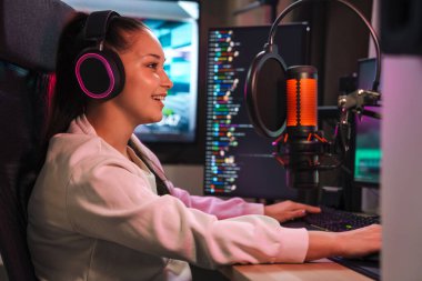 Evde bilgisayar başında kulaklık takan mutlu genç kadın oyuncu bilgisayar oyunları oynuyor.