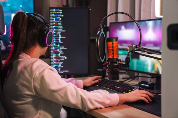 Back view of a young woman streamer gamer in headphones playing online games on pc computer at home