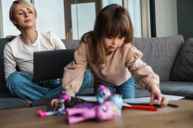 Genç anne ve anaokulu kızı oturma odasında, annesi dizüstü bilgisayarla çalışıyor.