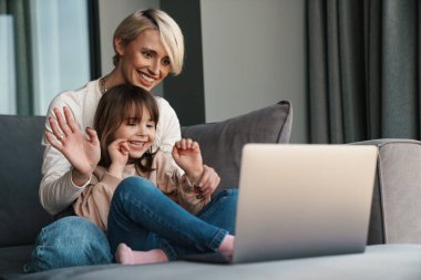 Mutlu genç anne ve küçük kızı bir kanepede dizüstü bilgisayarla oturuyor, ekrana bakıyor, video izliyor ya da film seyrediyor, el sallıyor.