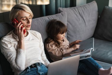 Mutlu genç anne ve küçük kızı bir kanepede dizüstü bilgisayarla oturuyor, ekrana bakıyor, video izliyor veya film seyrediyor, cep telefonuyla konuşan bir kadın.