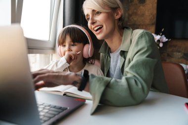 Mutlu anne kızına okul ödevlerinde yardım ediyor, dizüstü bilgisayar kullanıyor, karantina sırasında ders çalışıyor, kulaklıkla öğretmenini dinleyen küçük bir kız.