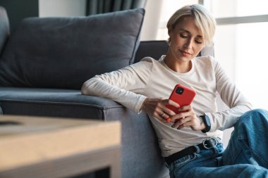 Evdeki koltukta cep telefonu kullanan orta yaşlı sarışın bir kadın.