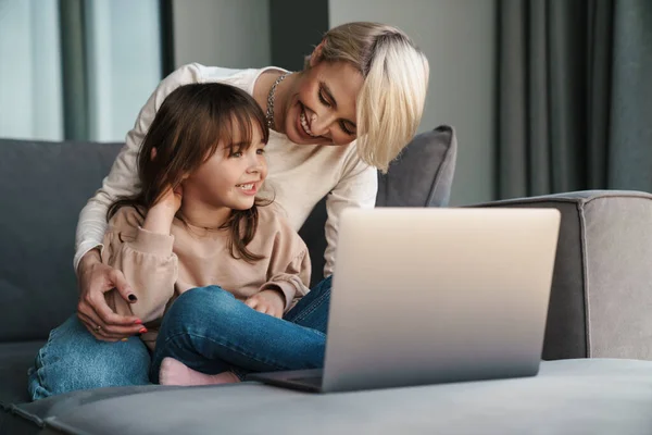 快乐的年轻妈妈和她的小女儿坐在沙发上 带着笔记本电脑 看着屏幕 打电话或看电影 — 图库照片