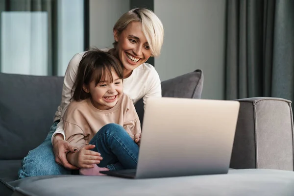快乐的年轻妈妈和她的小女儿坐在沙发上 带着笔记本电脑 看着屏幕 打电话或看电影 — 图库照片