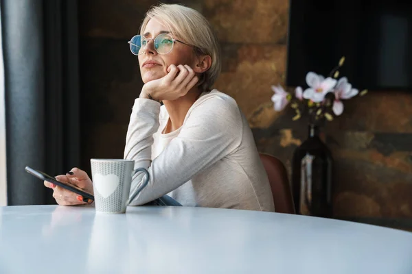 コーヒーを飲みながらキッチンテーブルに座っている間 携帯電話を使用してカジュアルな服を着て失望した若い女性を動揺 — ストック写真