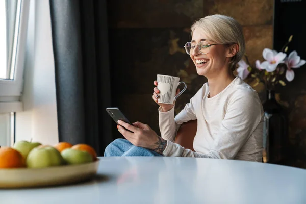 コーヒーを飲みながらキッチンテーブルに座りながら 携帯電話を使ってカジュアルな服装で笑顔の若い女性 — ストック写真