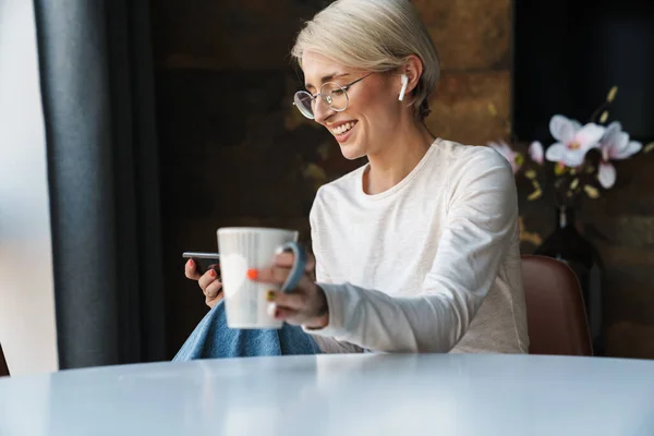 コーヒーを飲みながらキッチンテーブルに座りながら 携帯電話を使ってカジュアルな服装で笑顔の若い女性 — ストック写真
