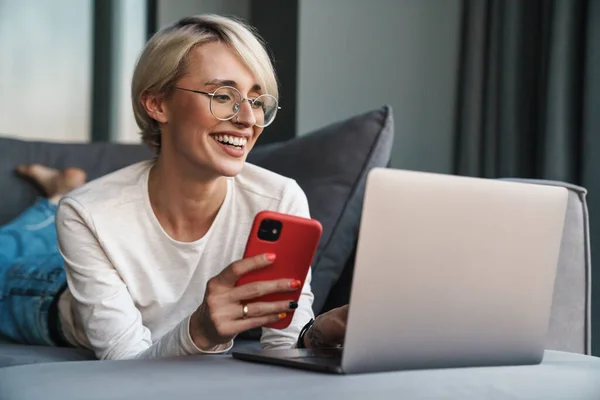 自宅のソファの上で携帯電話やラップトップコンピュータを使用して中年のブロンドの女性を笑顔 勉強や仕事 — ストック写真