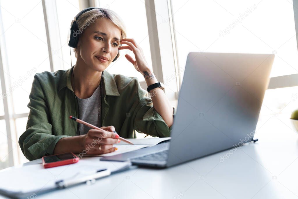 Smiling middle aged blonde woman with short hair studying or working on a video call via laptop computer while sitting at the table at home