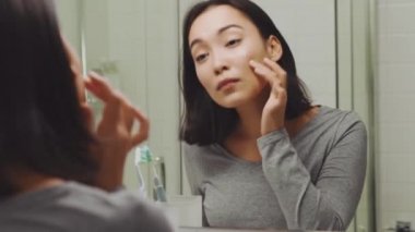 Yakışıklı Asyalı kadın aynada tenine bakıyor ve banyoda saçını tarıyor.