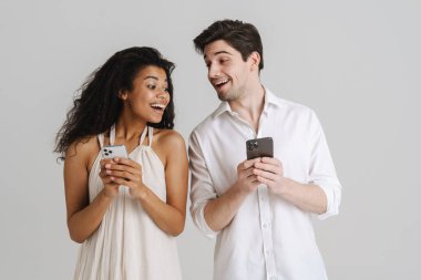 Beyaz arka planda izole edilmiş cep telefonlarını kullanırken gülümseyen çok ırklı genç çift.