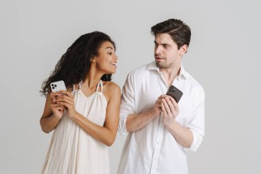 Beyaz arka planda izole edilmiş cep telefonları kullanan çok ırklı genç çift kaşlarını çatıyor