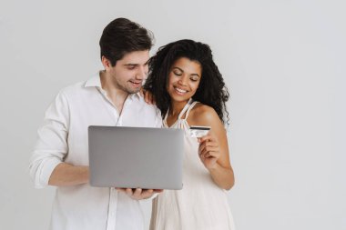 Laptop ve kredi kartıyla poz veren çok ırklı gülümseyen çift beyaz arka planda izole edilmiş.