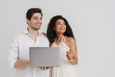 Laptop ve kredi kartıyla poz veren çok ırklı gülümseyen çift beyaz arka planda izole edilmiş.