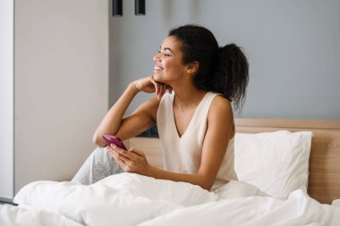 Yatakta elinde cep telefonu olan mutlu, Afrikalı bir kadın.