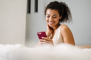 Yatakta elinde cep telefonu olan mutlu, Afrikalı bir kadın.