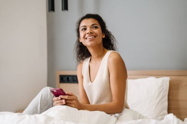 Yatakta elinde cep telefonu olan mutlu, Afrikalı bir kadın.