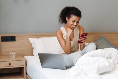 Genç siyah kadın, evde uyuduktan sonra yatakta otururken akıllı telefon ve laptop kullanıyor.