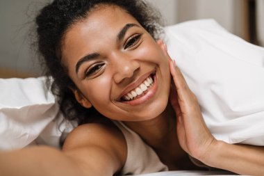 Mutlu, genç Afrikalı bir kadın battaniyenin altına saklanıp evde bir yatakta gülüyor ve selfie çekiyor.