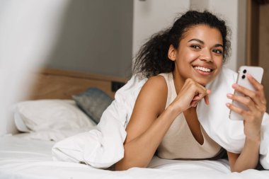 Genç siyahi kadın evde uyuduktan sonra yatakta yatarken gülümsüyor ve akıllı telefon kullanıyor.