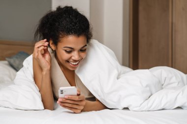 Genç siyahi kadın evde uyuduktan sonra yatakta yatarken gülümsüyor ve akıllı telefon kullanıyor.