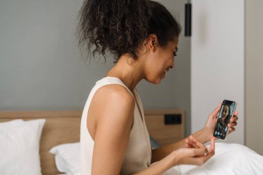 Mutlu, genç Afrikalı bir kadın yatak odasında oturmuş video görüşmesinde cep telefonunu tutuyor.