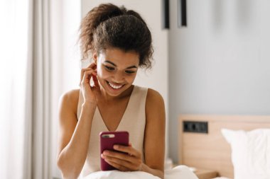 Yatakta elinde cep telefonu olan mutlu, Afrikalı bir kadın.