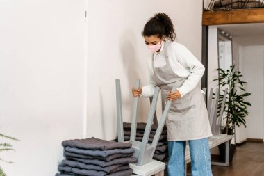 Kapalı kafede çalışan yüz maskesi ve önlük takan genç zenci garson.