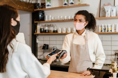 Yüz maskeli genç siyahi garson kapalı kafedeki ödeme terminalinde çalışıyor.