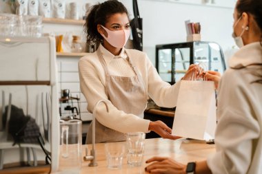 Yüz maskesi takan genç siyahi garson müşteriyle kapalı bir kafede çalışıyor.