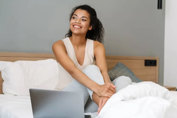 年轻的黑人妇女在家里睡觉后坐在床上使用笔记本电脑 — 图库照片