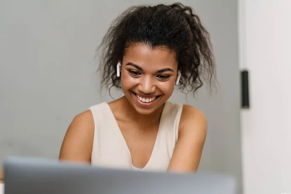快乐的年轻非洲女人坐在卧室的床上 戴着耳机 通过笔记本电脑通过视频通话 — 图库照片
