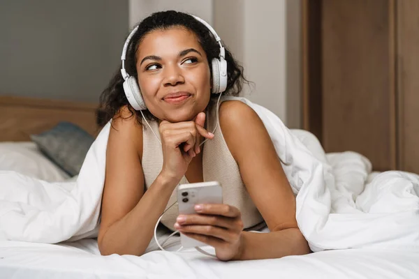 年轻的黑人妇女在家里睡觉后躺在床上用耳机和智能手机 — 图库照片