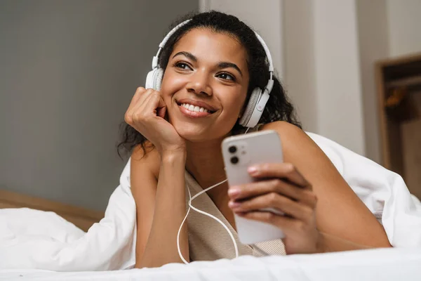 年轻的黑人妇女在家里睡觉后躺在床上用耳机和智能手机 — 图库照片