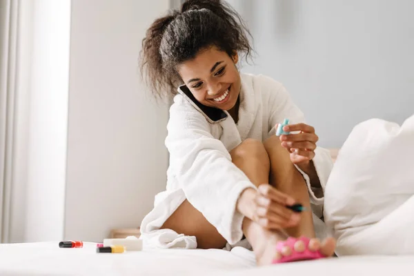 Sonriente Joven Africana Albornoz Sentada Una Cama Pintándose Las Uñas — Foto de Stock