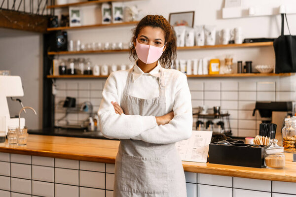Young black waitress in face mask standing by cafe counter indoors
