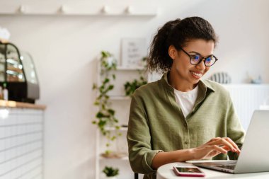 Gözlüklü genç siyahi kadın kafede otururken dizüstü bilgisayarla çalışıyor.