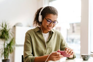 Kapalı kafede cep telefonu ve kulaklık kullanan genç siyahi kadın gülümsüyor.