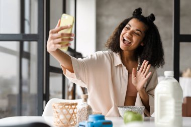 Evde kahvaltı ederken cep telefonundan selfie çeken genç siyahi bir kadın.