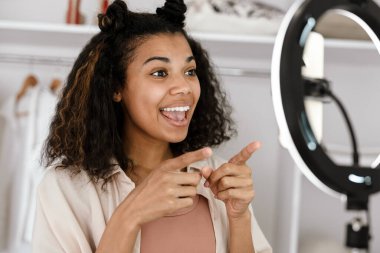 Siyah genç bir kadın kostüm dolabında cep telefonundan selfie çekerken el kol hareketi yapıyor.