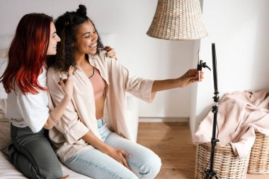 İki mutlu, etnik çeşitliliğe sahip genç bayan arkadaş, kameranın önünde yatakta oturmuş, yaşam tarzı videosu çekiyorlar.