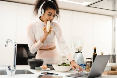 Kulaklıklı siyahi kadın evde smoothie yaparken dizüstü bilgisayar kullanıyor.