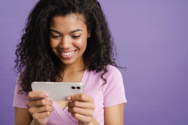 Mor arka planda izole edilmiş, gülen ve cep telefonu kullanan genç siyah bir kadın.