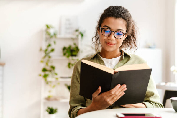 Young black woman in eyeglasses reading book while sitting at cafe indoors