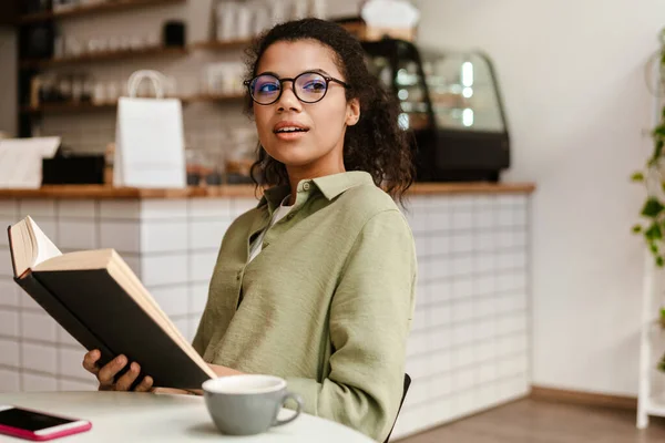 Młoda Czarna Kobieta Okularach Czyta Książkę Podczas Picia Kawy Kawiarni — Zdjęcie stockowe