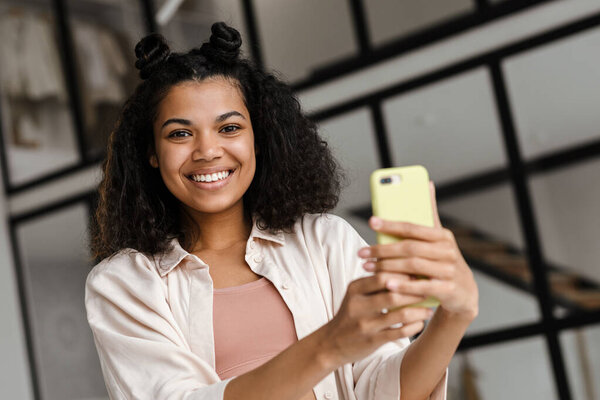 Black young woman smiling while taking selfie on mobile phone at home