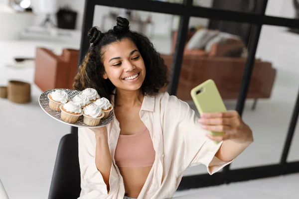 Happy young african woman blogger making a video on mobile phone holding plate with cupcake standing in the room