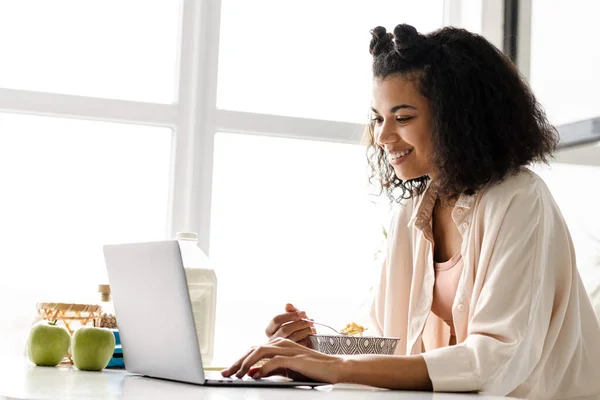 年轻黑人妇女在家里吃早餐的时候带着笔记本电脑工作 — 图库照片